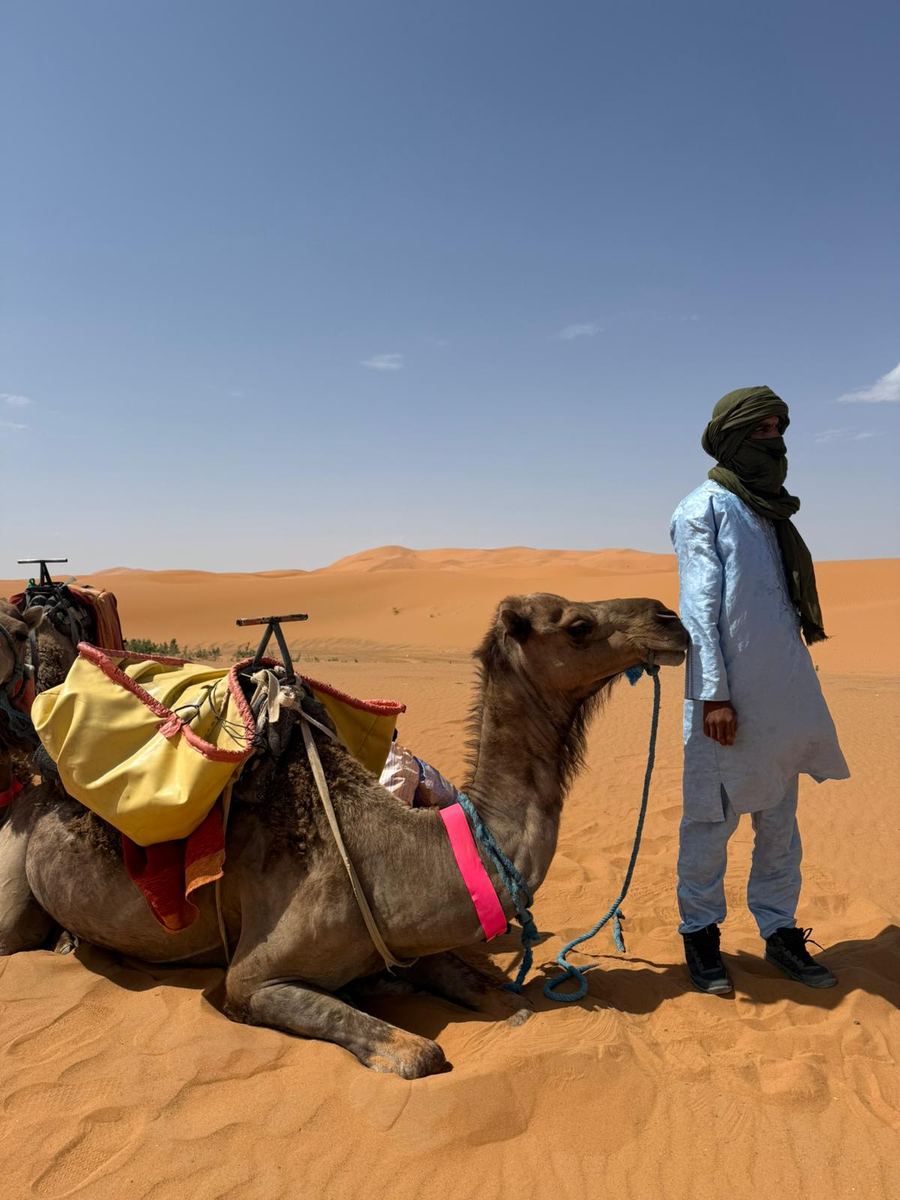 Two travelers in desert headscarves standing beside a resting camel with their Berber guide in the Sahara